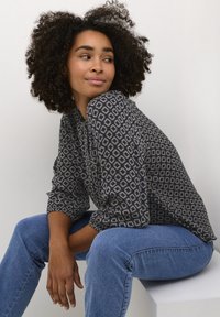 Musterbluse in Marineblau mit cremefarbenen geometrischen Rauten, langen Ärmeln und lockerer Passform. Kombiniert mit hellblauen Jeans.