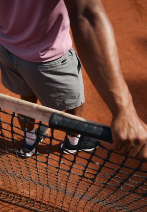 Tenue de sport comprenant un t-shirt rose clair et un short gris. Détails notables : logo de la marque sur le short, chaussures de tennis noires et une raquette de tennis.