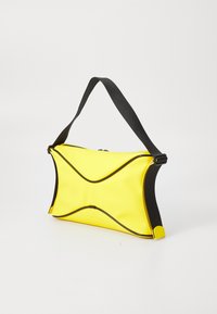 Gul veske med blank overflate, med bølgete design, svarte detaljer og en kontraststropp. Glidelås lukking på toppen.