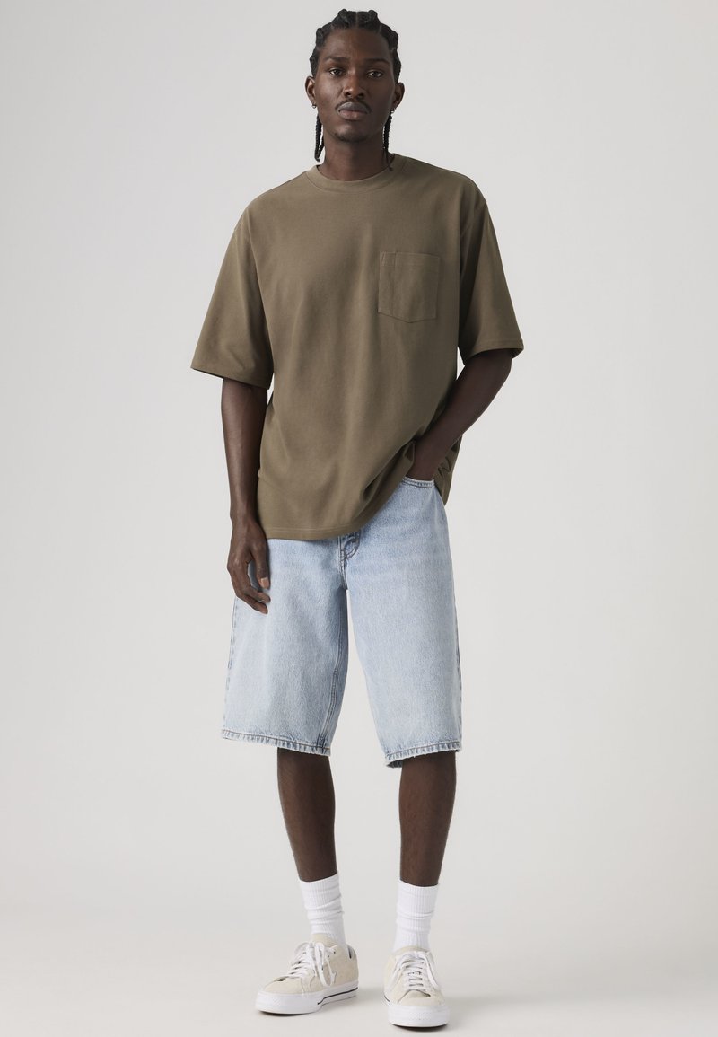 Mann steht mit der Hand in der Tasche seiner hellblauen Jeansshorts, trägt ein olivgrünes übergroßes T-Shirt, weiße Socken und beige Turnschuhe.