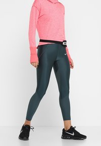 Completo sportivo composto da una felpa con cappuccio a maniche lunghe di un vivace colore rosa, Leggings verde scuro con pannelli testurizzati e scarpe da ginnastica nere con il logo argentato.