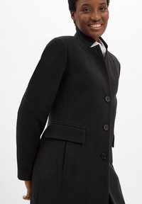 Cappotto di lana nera con colletto alto, dotato di due tasche laterali, cinque bottoni neri e spalle strutturate. Texture liscia, vestibilità sartoriale.