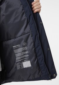 Vnitřek námořnické bundy se hladkým, lesklým povrchem; obsahuje štítek s informacemi o účincích větrného chladu, pokyny k údržbě a teplotními doporučeními.