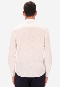 Uomo con capelli corti e scuri, che indossa una camicia di lino bianca a maniche lunghe e pantaloni scuri, in piedi con la schiena rivolta verso uno sfondo bianco.