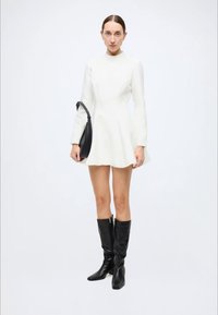 White long-sleeve dress with a flared skirt, high neck, and smooth texture, paired with knee-high black boots and a shoulder bag.