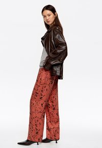 Chaqueta de cuero marrón sobre una camiseta gris, combinada con pantalones de pierna ancha en color coral con un patrón de salpicaduras negras. Zapatos negros de punta completan el conjunto.