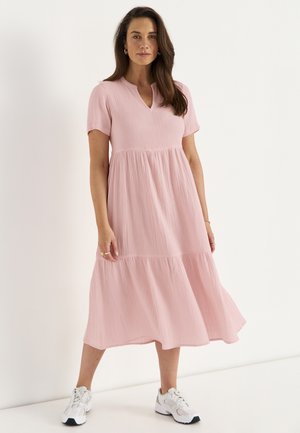 Nainen pukeutuneena soljuvaan vaaleanpunaiseen midimekkoon, jossa on lyhyet hihat ja valkoiset tennarit, seisoo neutraalia taustaa vasten.