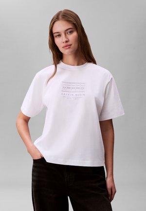 Jeune femme aux longs cheveux portant un t-shirt blanc Calvin Klein avec un croquis de bâtiment et un pantalon noir, debout devant un fond gris.