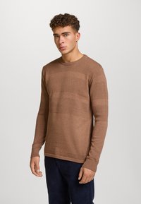 Maglione marrone lavorato a maglia con motivi a righe testurizzate, collo rotondo, maniche lunghe e polsini a coste. Indossato con pantaloni scuri.