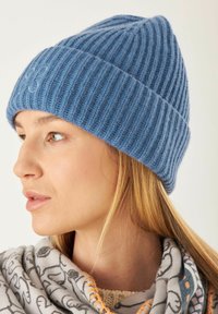 Blauwe gebreide beanie met geribbelde textuur, voorzien van een klein logo detail aan de voorkant, gedragen met een gebreide sjaal.