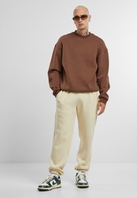 Brun oversized sweatshirt med ribbet halsudskæring, cremefarvede tapered sweatpants og grønne og hvide sneakers med en struktureret overflade.
