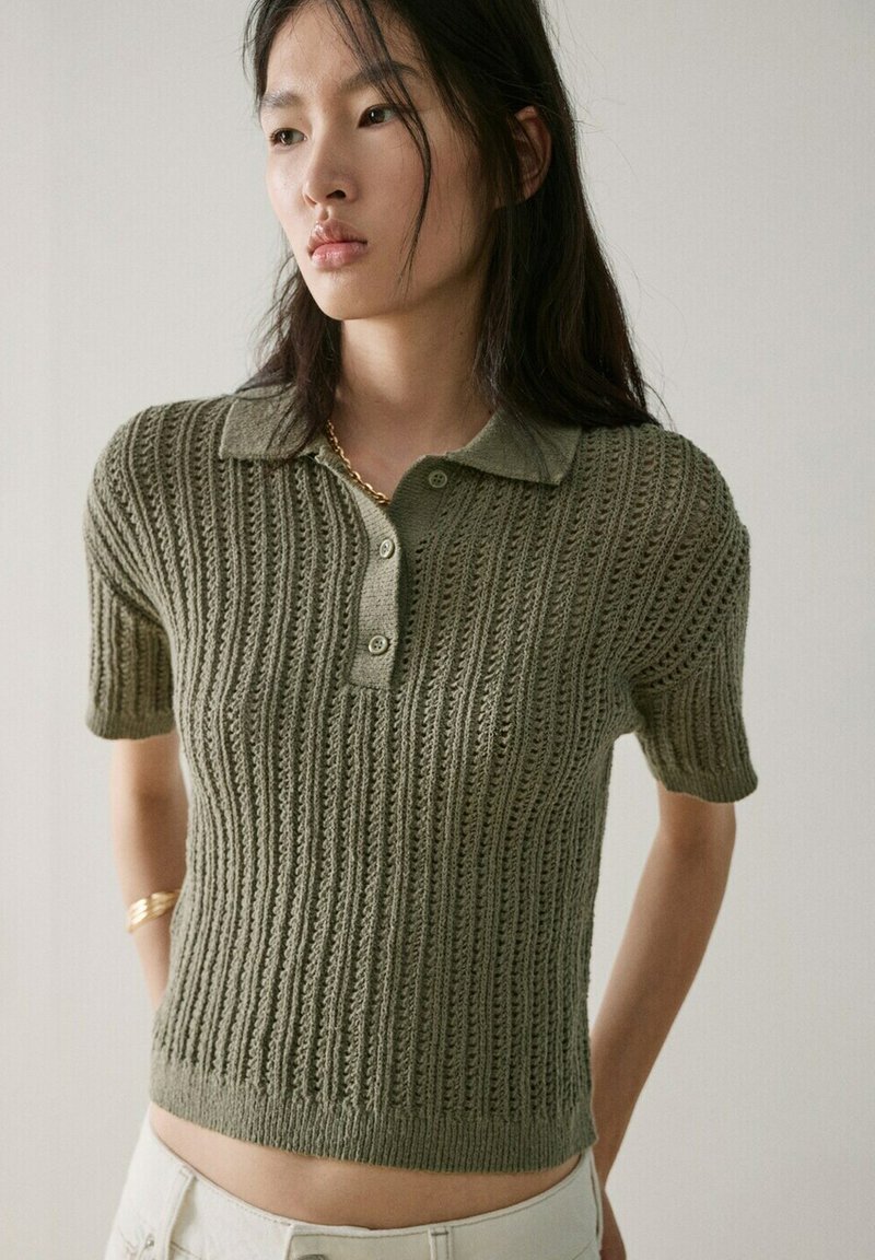 Jeune femme portant un polo en maille à manches courtes vert olive avec un motif texturé, un collier en or et un bracelet en or, debout devant un fond uni.
