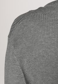 Grå stickad tröja med ribbat mönster; har en bred krage och en subtil glans. Materialet verkar mjukt och stretchigt, med en figurnära design.