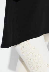 Top nero con orlo arrotondato abbinato a pantaloni color crema con ricami floreali intricati e dettagli a taglio lungo il lato.