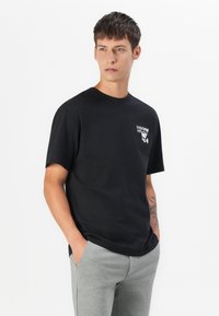Camiseta de algodón negra con mangas cortas y escote redondo, con un gráfico de logotipo en la parte frontal en blanco. Llevada con pantalones de chándal gris claro.