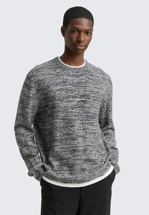 Jeune homme portant un pull gris texturé à manches longues par-dessus une chemise blanche, les mains dans les poches, devant un fond clair uni.