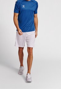 Modré sportovní tričko s krátkým rukávem, kombinované s bílými šortkami. Tričko má na hrudi logo a hladkou texturu. Bílé tenisky.