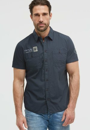 Mann mit kurzen braunen Haaren, der ein dunkel marineblaues kurzärmeliges Hemd mit Knöpfen und hellblaue Jeans trägt, vor einem schlichten Hintergrund steht.