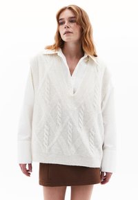 Gilet en tricot blanc surdimensionné avec un col en V, présentant un motif de câble texturé. Porté par-dessus une chemise blanche à boutons et une jupe marron.