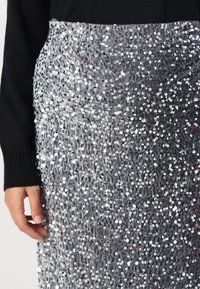 Gonna argento in paillettes con superficie testurizzata, dotata di vita aderente e una mano che poggia sul tessuto. Lo sfondo è neutro.