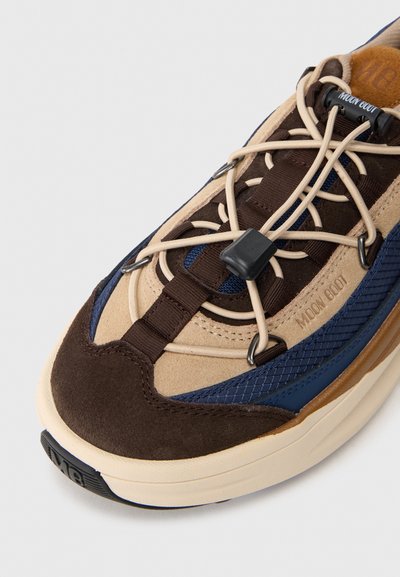 Zbliżenie brązowego, beżowego i granatowego sneaker'a Moon Boot z beżowymi sznurkami i teksturowaną gumową podeszwą na białym tle.