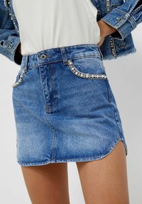 Jeansowa spódnica mini z perłowymi i dżetowymi zdobieniami na przednich kieszeniach, zestawiona z białą bluzką oraz dżinsową kurtką z podobnymi ozdobami.