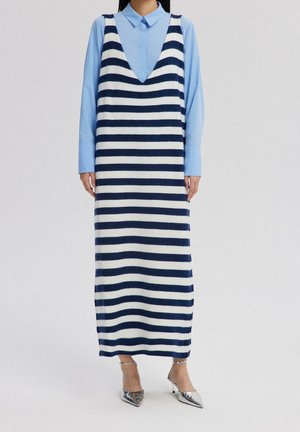 Femme portant une robe en tricot sans manches rayée bleu marine et blanc par-dessus une chemise bleu clair à col, avec des talons pointus argentés.
