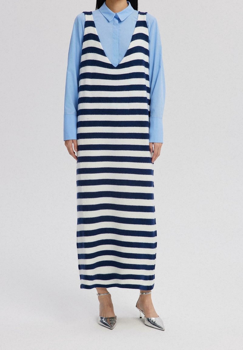 Femme portant une robe en tricot sans manches rayée bleu marine et blanc par-dessus une chemise bleu clair à col, avec des talons pointus argentés.