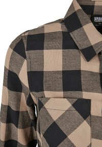 Chemise à carreaux beige et noire, dotée d'un col, de manches longues et d'une poche avant avec un tissu texturé et un motif tartan distinct.