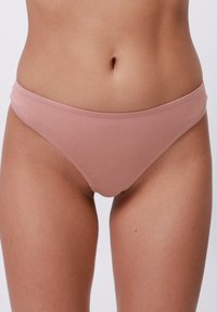 Egy nő világos rózsaszín pamut bikini stílusú fehérneműt visel, amely a has alsó részét és a combok felső részét mutatja, fehér háttér előtt.