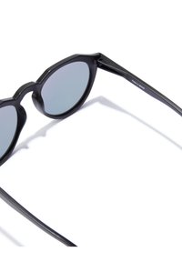 Musta aurinkolasit pyöreillä kehillä, harmaat linssit ja mattapinta. Varsissa on tyylikäs hieman kaareva muoto, merkitty "Valmistettu Espanjassa."