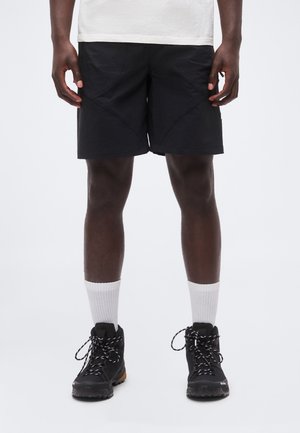 Personne portant un short texturé noir, des chaussettes blanches montantes et des chaussures de randonnée noires avec des lacets noirs et blancs sur fond blanc.