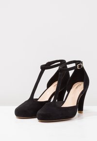 Mustat mokkanahkasaappaat, joissa on pyöreä kärki, T-nauhatyyli ja kultasävyinen solkiyksityiskohta. Korko on noin 10 cm korkea.