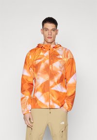 Orange mønstrede vindjakke med hætte, der har en lynlåslomme, letvægtsmateriale og elastiske manchetter.