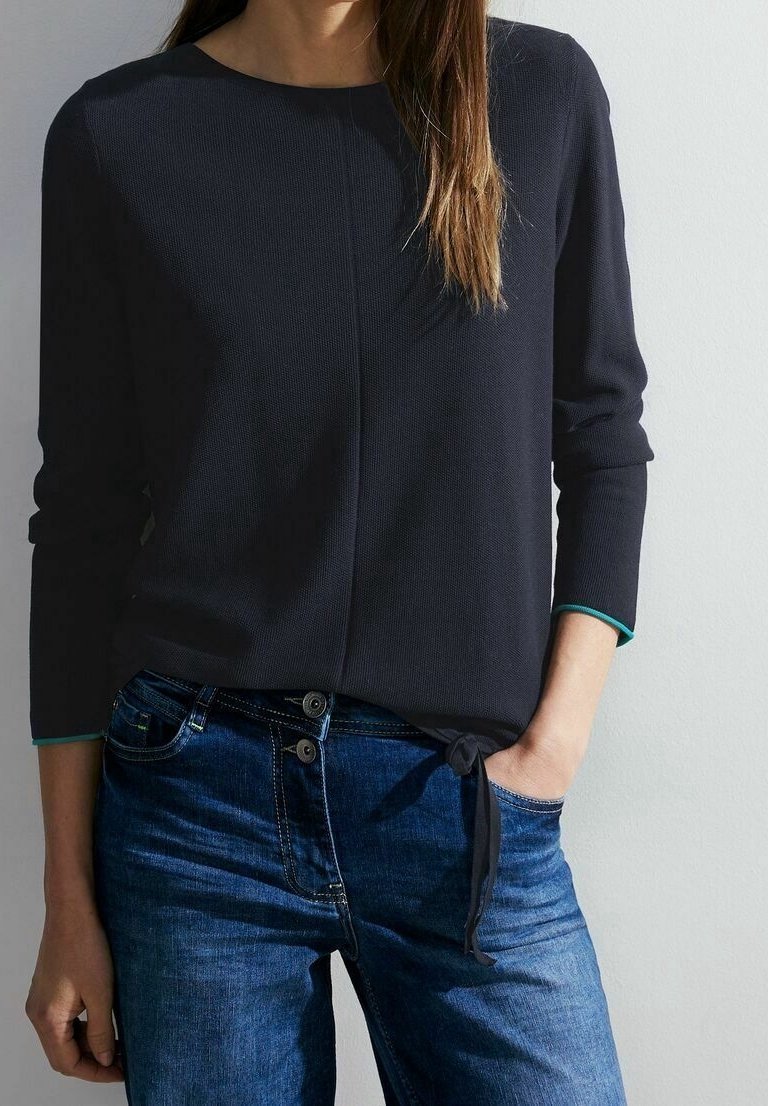 Femme portant un haut à manches longues foncé avec un détail noué sur le côté et un jean bleu, debout avec une main dans la poche devant un fond clair.