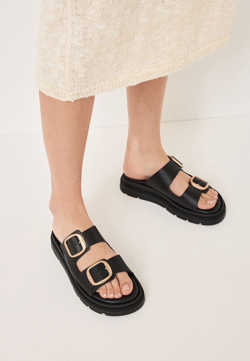 Zwarte slipper-sandalen met twee brede banden met vierkante gouden gespen, dikke zool met een gestructureerde grip, en een open teen ontwerp.