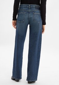 Weite blaue Denim-Jeans mit glatter Textur, ausgestattet mit hinteren Taschen, die mit silbernen Nieten verziert sind, und einem hohen Bund.