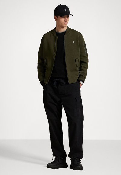 Blusão bomber verde-oliva por cima de uma t-shirt preta com gola redonda, combinado com calças cargo largas pretas e ténis pretos. Boné preto completa o visual.