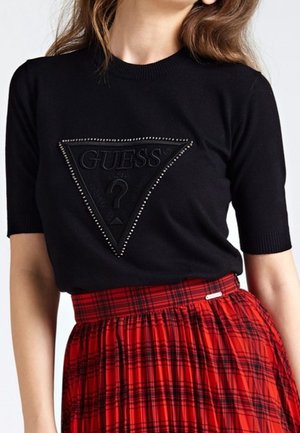 Czarny top z krótkim rękawem z ćwiekowanym trójkątnym logo Guess, zestawiony z czerwoną i czarną kratowaną spódnicą z wysokim stanem.