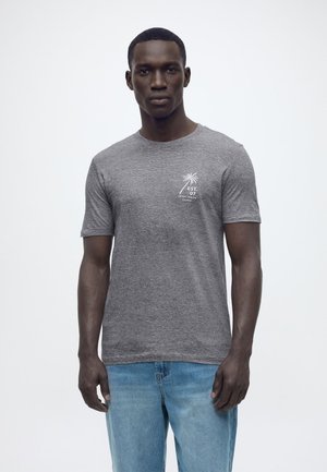 Άνδρας που φορά γκρι κοντομάνικη μπλούζα με ένα μικρό λευκό γραφικό φοίνικα και κείμενο στο αριστερό στήθος, συνδυασμένη με ανοιχτόχρωμα τζιν.