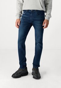 Dark blue denim jeans with a fitted design, featuring front pockets and a button closure, paired with textured black athletic shoes.