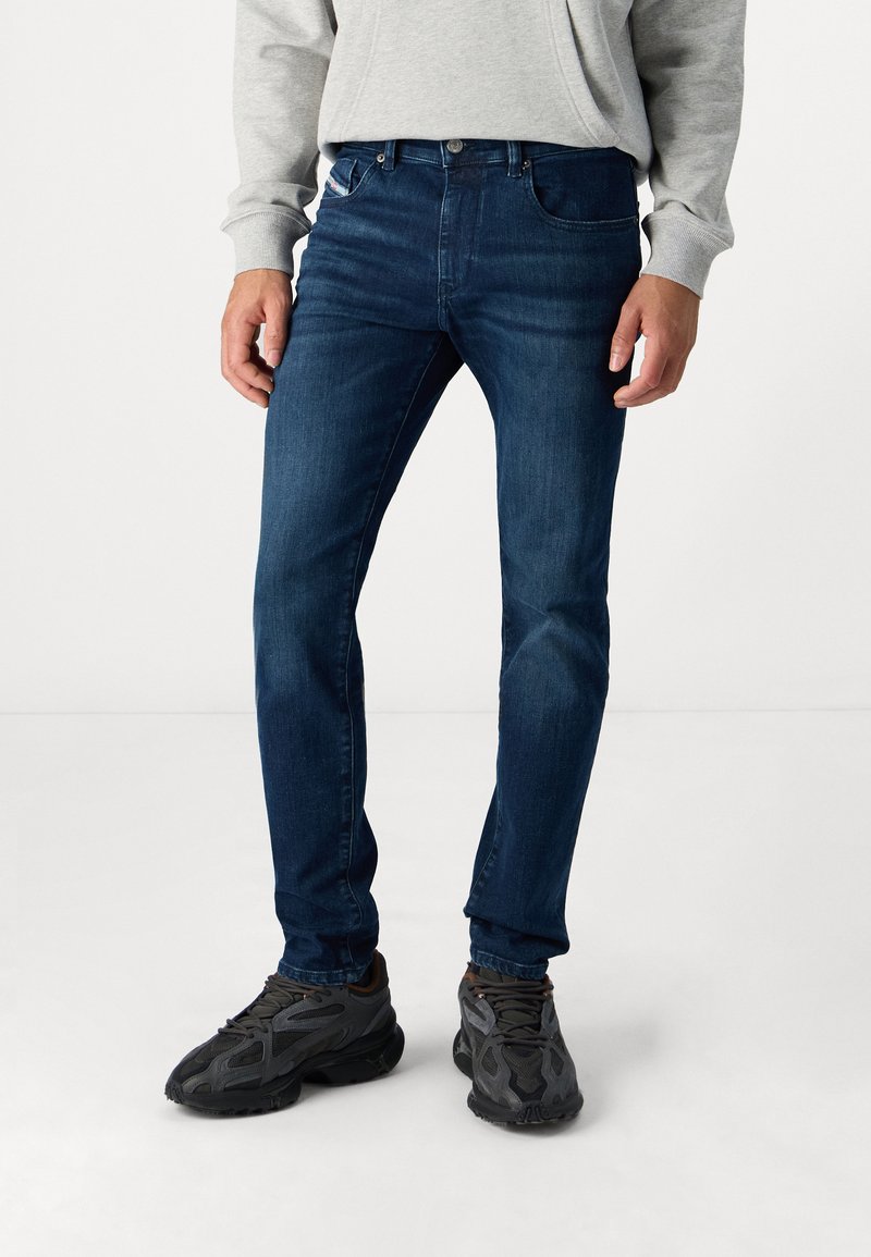 Dark blue denim jeans with a fitted design, featuring front pockets and a button closure, paired with textured black athletic shoes.