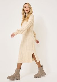 Knit cable sweater dress in cream with a relaxed fit, long sleeves, side slits, and ribbed edges, paired with taupe ankle boots.