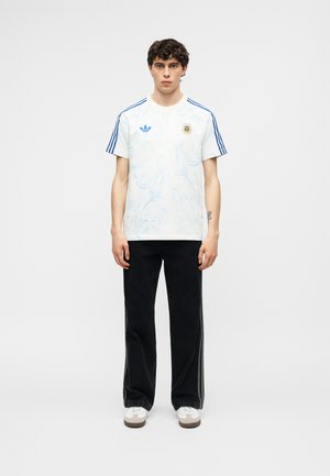 Man draagt een wit Argentijns voetbalshirt met blauwe strepen en zwarte broek, staand tegen een effen lichtgrijze achtergrond.