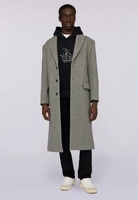 Szary długi płaszcz z wełny z klapami z wcięciem i dwoma przednimi kieszeniami, noszony na czarnej bluzie z kapturem, zestawiony z ciemnymi spodniami i białymi tenisówkami.