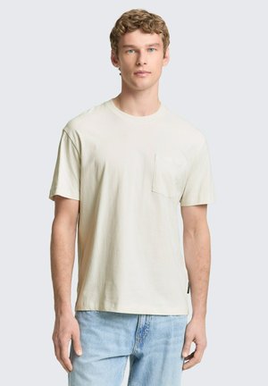 Młody mężczyzna o kręconych blond włosach, ubrany w gładki, kremowobiały, krótki rękaw z kieszenią na piersi oraz jasnoniebieskie dżinsy.