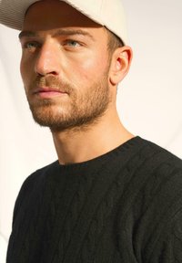 Czarny sweter z warkoczykovym splotem i okrągłym dekoltem, w zestawie z jasną czapką. Materiał wydaje się być strukturalny i ciepły.