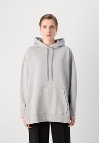 Hanorac oversized gri, realizat dintr-un material moale, cu o buzunar tip kangaroo, glugă cu șnur ajustabil și manșete ribbate, purtat peste pantaloni negri.