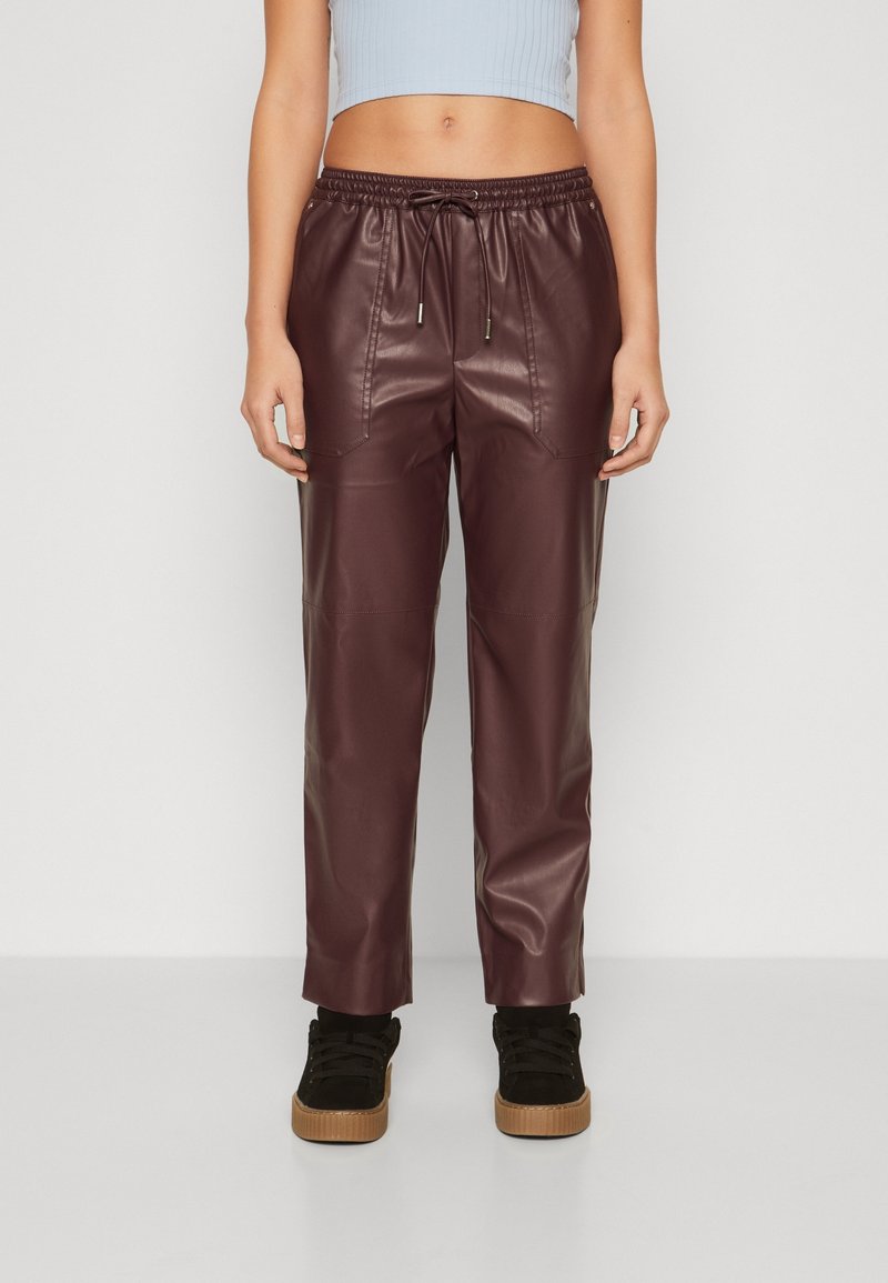 Femme portant un pantalon en simili cuir marron avec des poches avant, associé à des baskets noires et un top court bleu clair, debout sur un sol blanc.