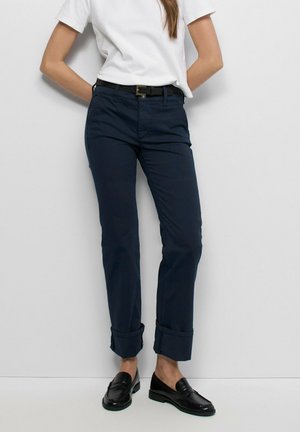 Persona que lleva pantalones azul oscuro con puños, una camisa blanca de manga corta, cinturón negro y mocasines negros, de pie contra una pared blanca.